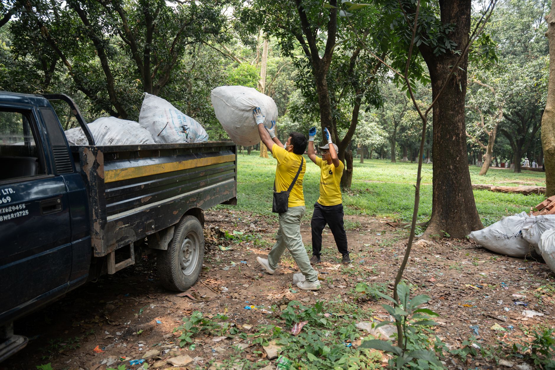 Two workers in yellow shirts load a truck with large garbage bags in a wooded park area.