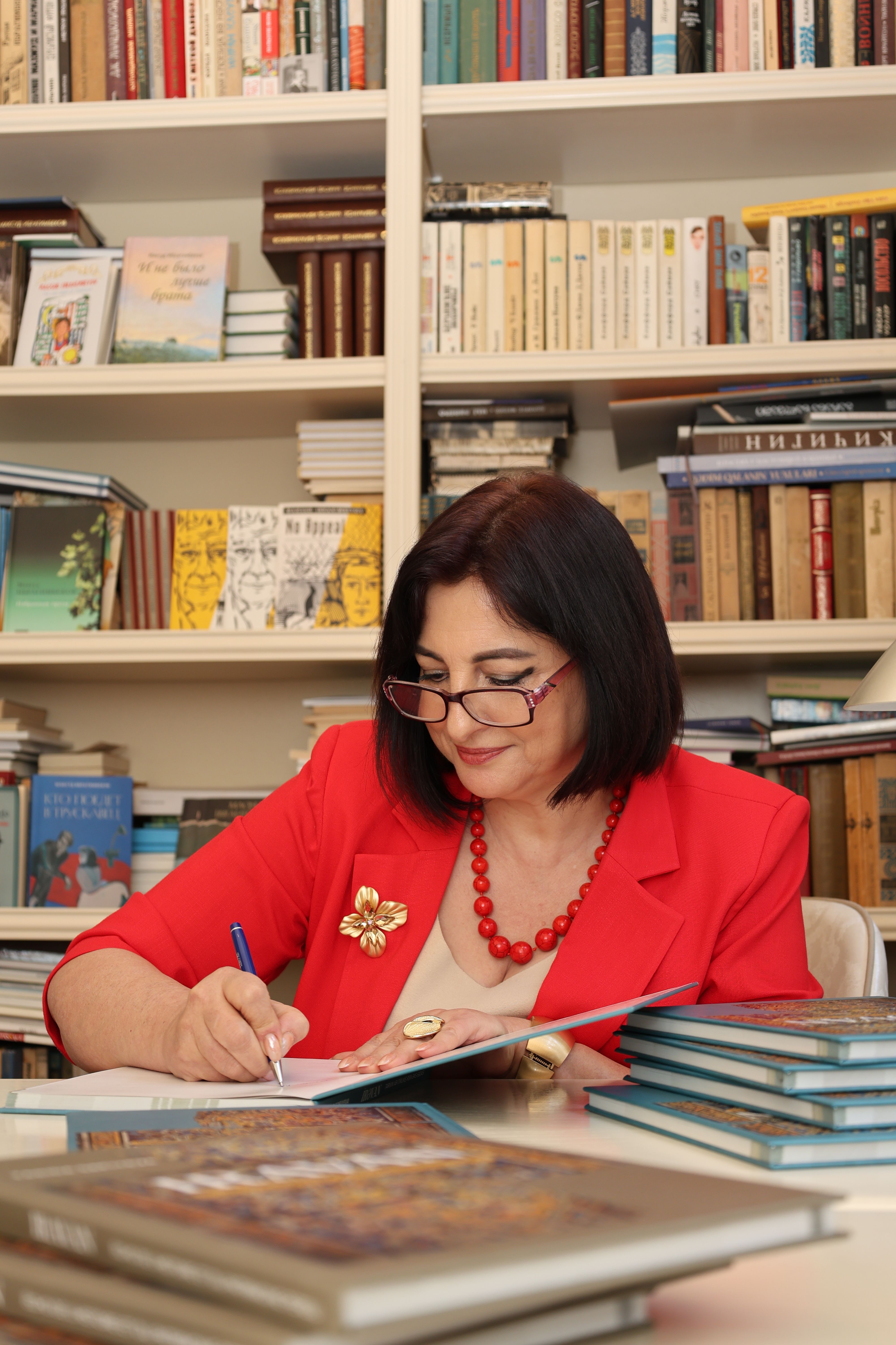 Adult woman writing in a library surrounded by books, wearing red.