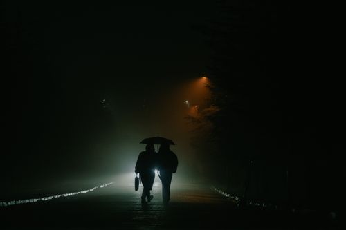 Free Moody silhouette of a couple with umbrella strolling under street lights in a foggy street. Stock Photo