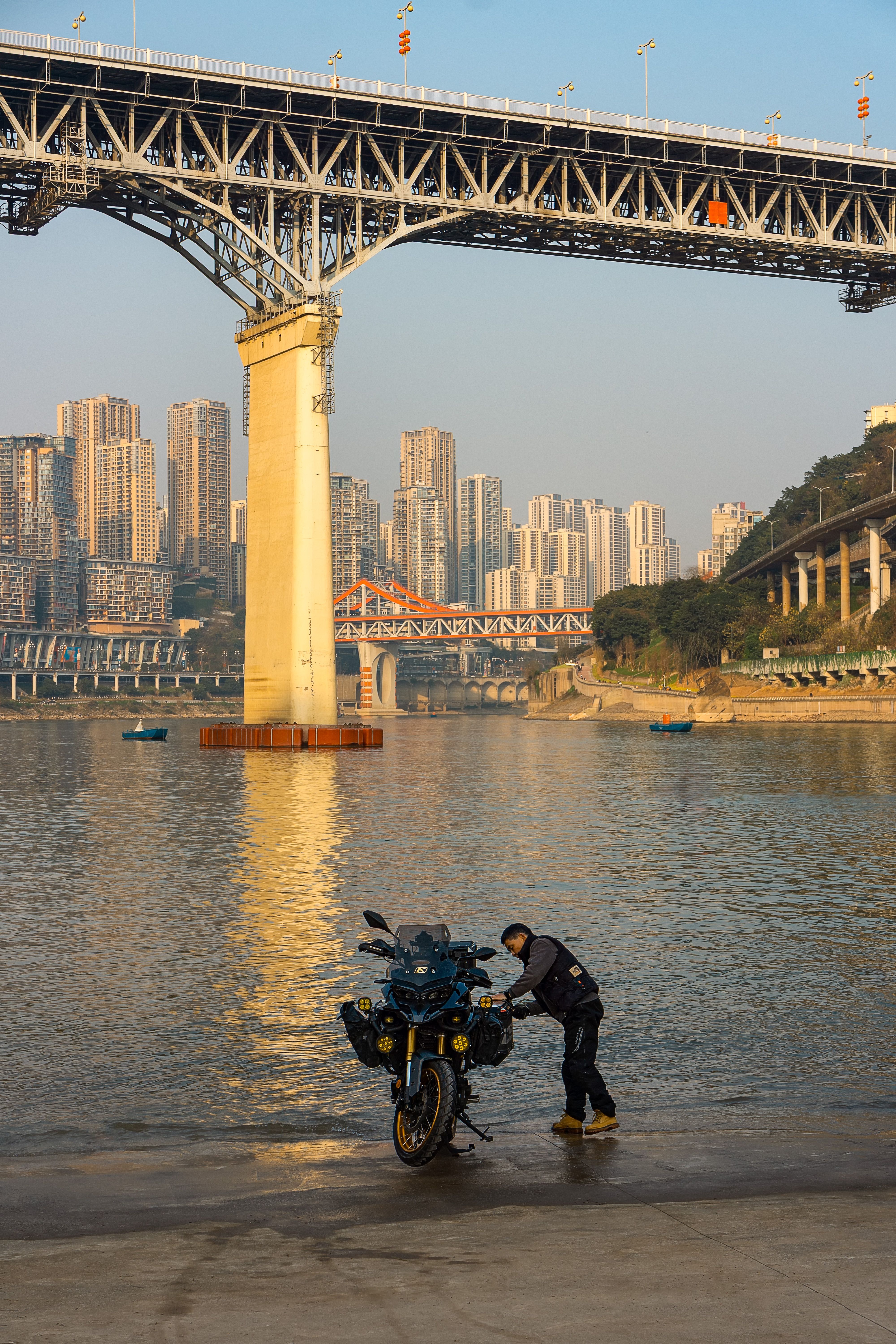 Motorcyclist beside bike under city bridge with skyline in the background at sunset.