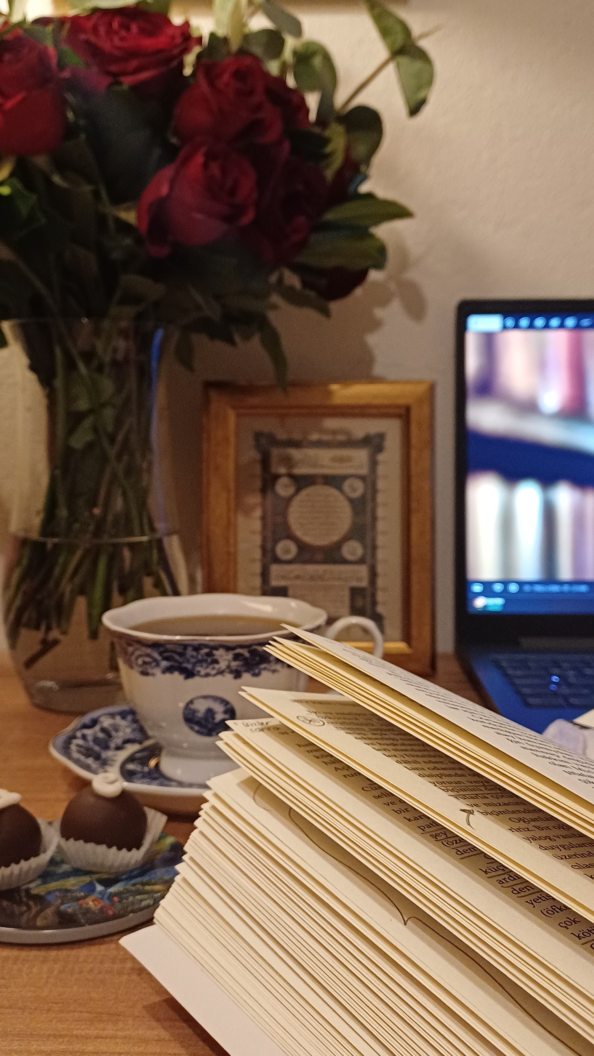 A cozy reading setup with a book, tea, laptop and roses, creating a perfect atmosphere for relaxation at home.