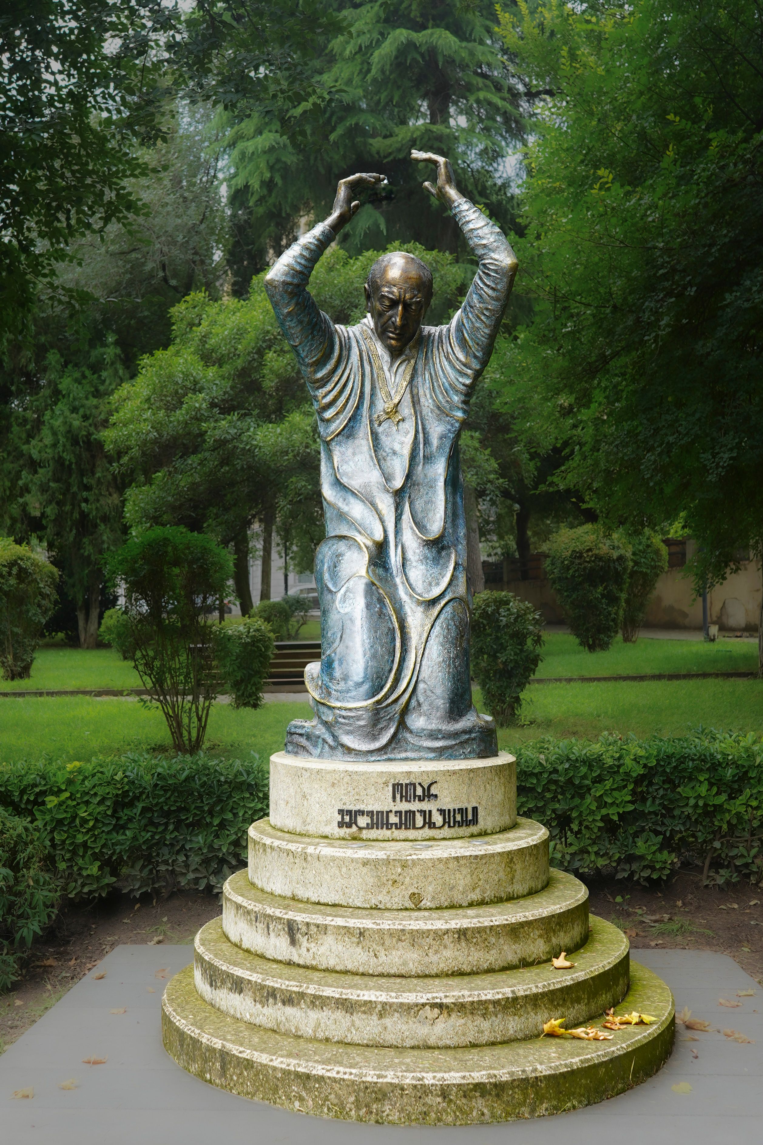 Bronze statue of a man with arms raised amid lush green trees, showcasing artistic sculpture.