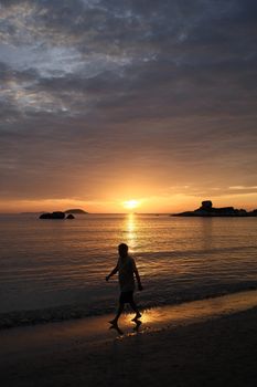 Silhouette of a person walking on the beach during a stunning sunset over calm ocean waters.