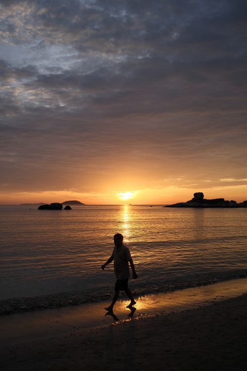Free Silhouette of a person walking on the beach during a stunning sunset over calm ocean waters. Stock Photo