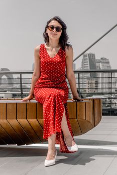 Fashionable woman in red polka dot dress sitting outside in urban setting.