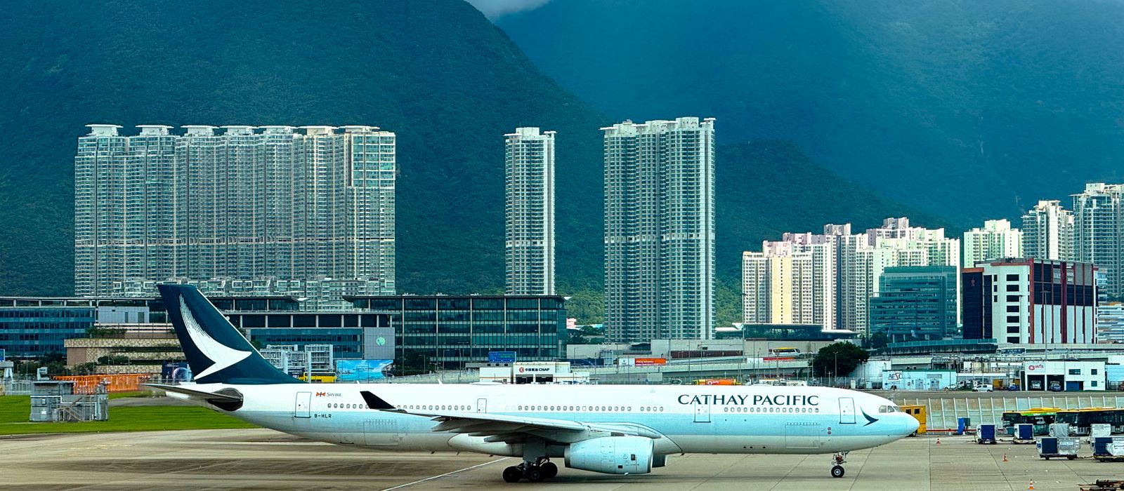 Cathay Pacific airplane taxiing on runway with Hong Kong skyline and mountains in the background.