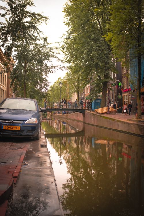 Free A picturesque canal view in Amsterdam with a car parked nearby and people walking along the street. Stock Photo