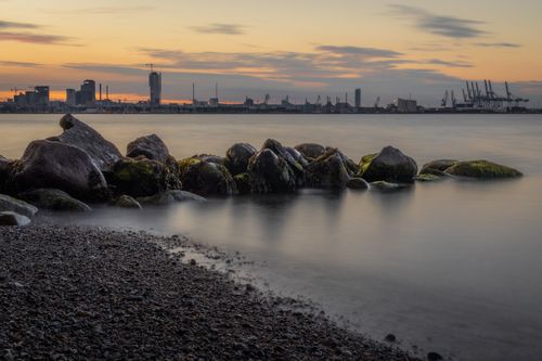 Free Serene coastal view with industrial skyline at twilight, showcasing calm waters and rocks. Stock Photo