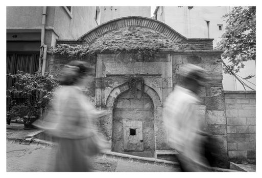 Blurred pedestrians pass a historic archway in İstanbul, Türkiye, in black and white.