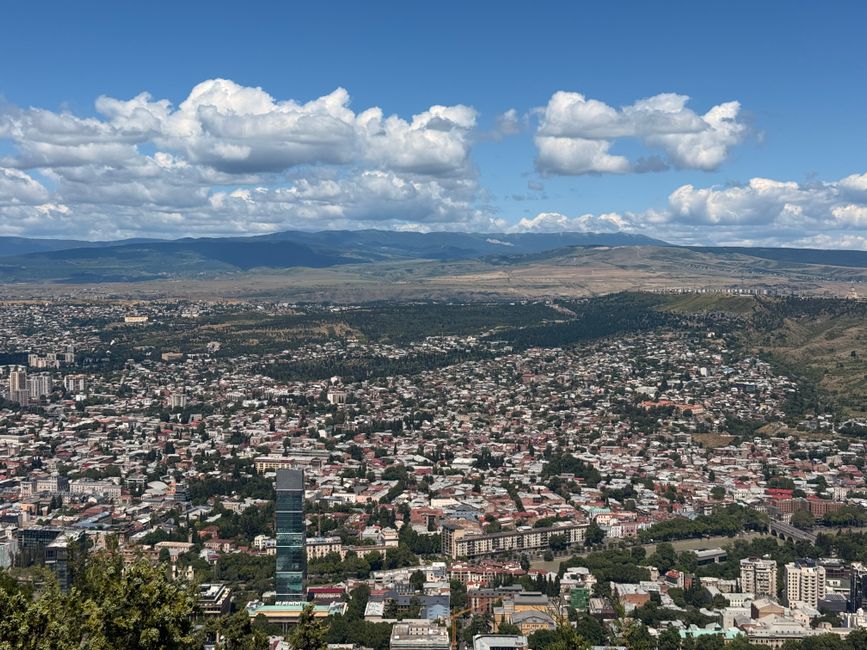 Aerial view of Tbilisi, Georgia, showcasing cityscape against mountain backdrop under a blue sky.