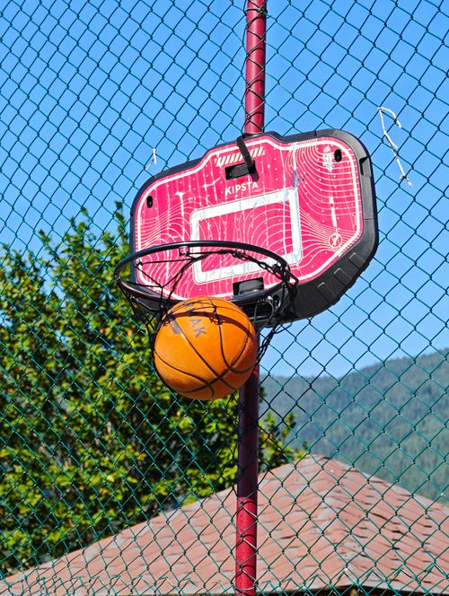 Free A basketball stuck in a red outdoor hoop against a clear blue sky and greenery. Stock Photo