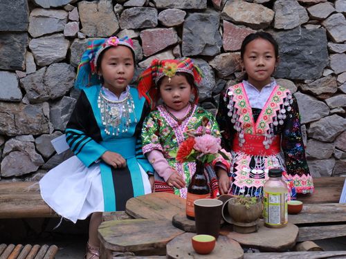 Free Three young girls in colorful traditional Hmong clothing sit on a wooden bench against a stone wall. Stock Photo