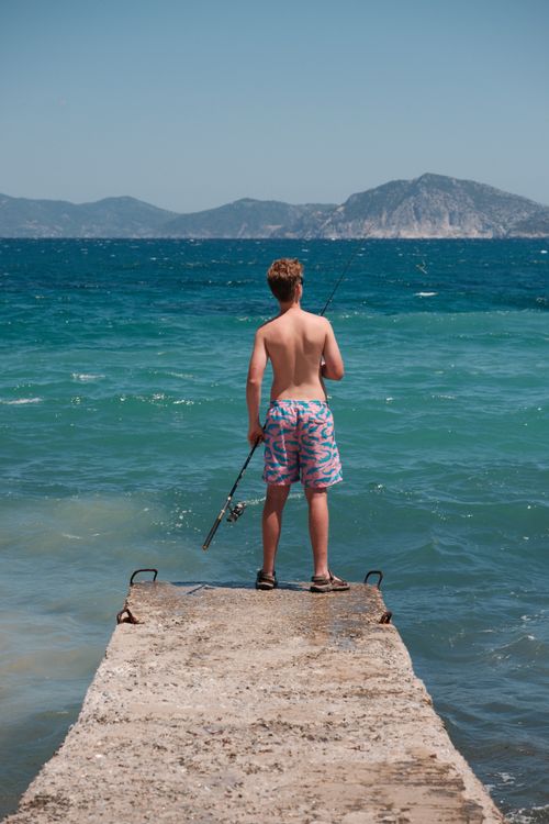 Free A teenager enjoys fishing from a concrete pier on a sunny day with a beautiful ocean backdrop. Stock Photo