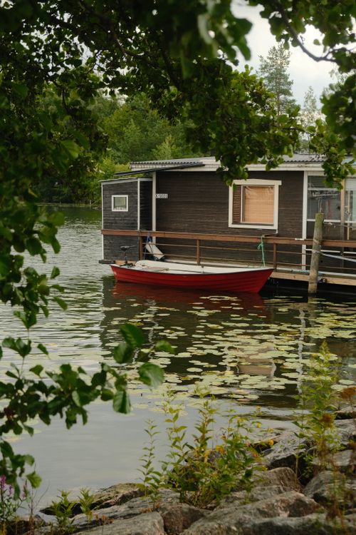 Free Serene lakeside scene with a red boat beside a rustic houseboat surrounded by nature. Stock Photo