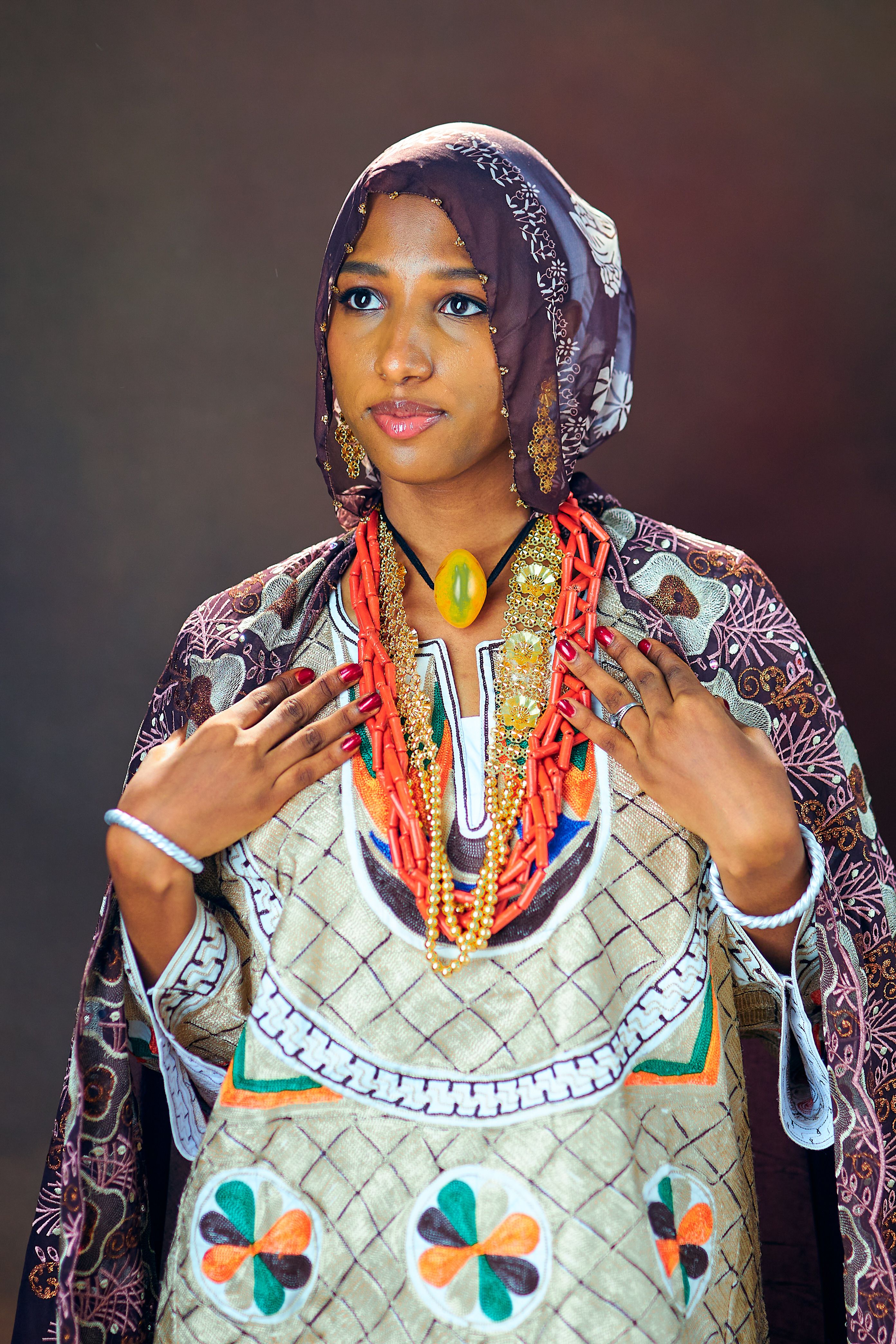 A woman in vibrant Nigerian traditional clothing and jewelry poses gracefully against a studio backdrop.