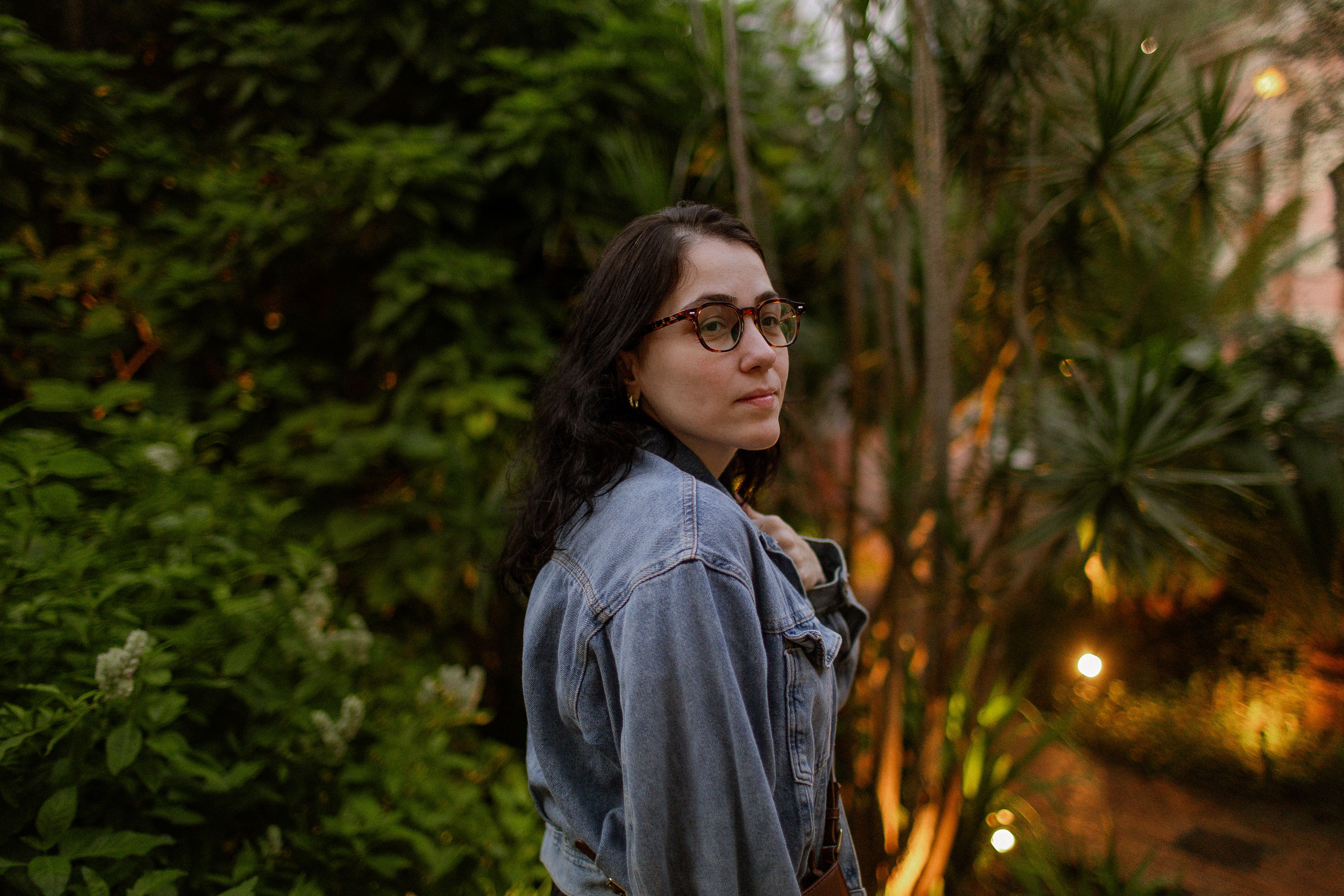 Young woman in denim jacket and glasses standing in a lush garden at dusk.