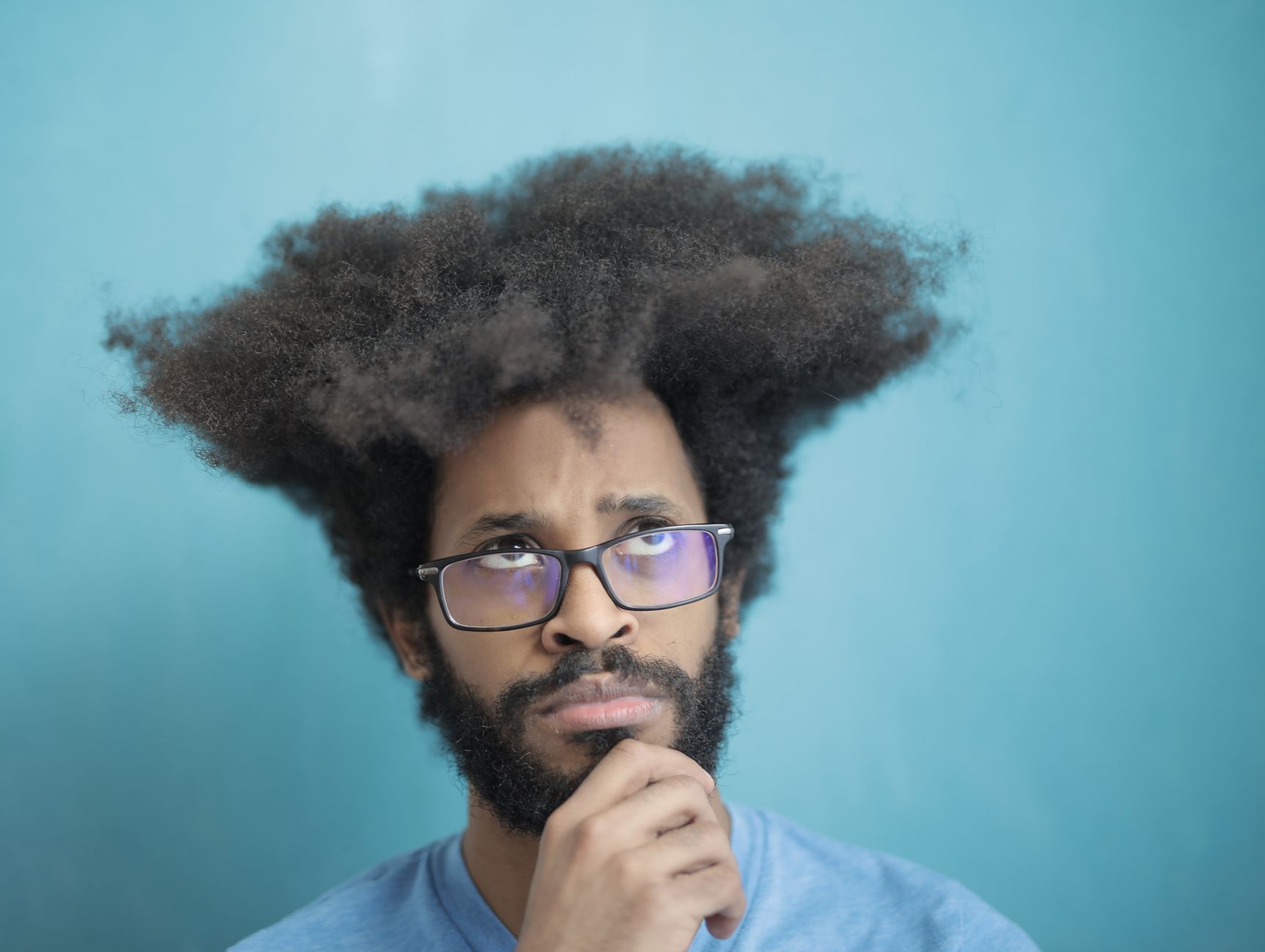 Portrait of a thoughtful man with a unique afro hairstyle, wearing glasses against a blue backdrop.
