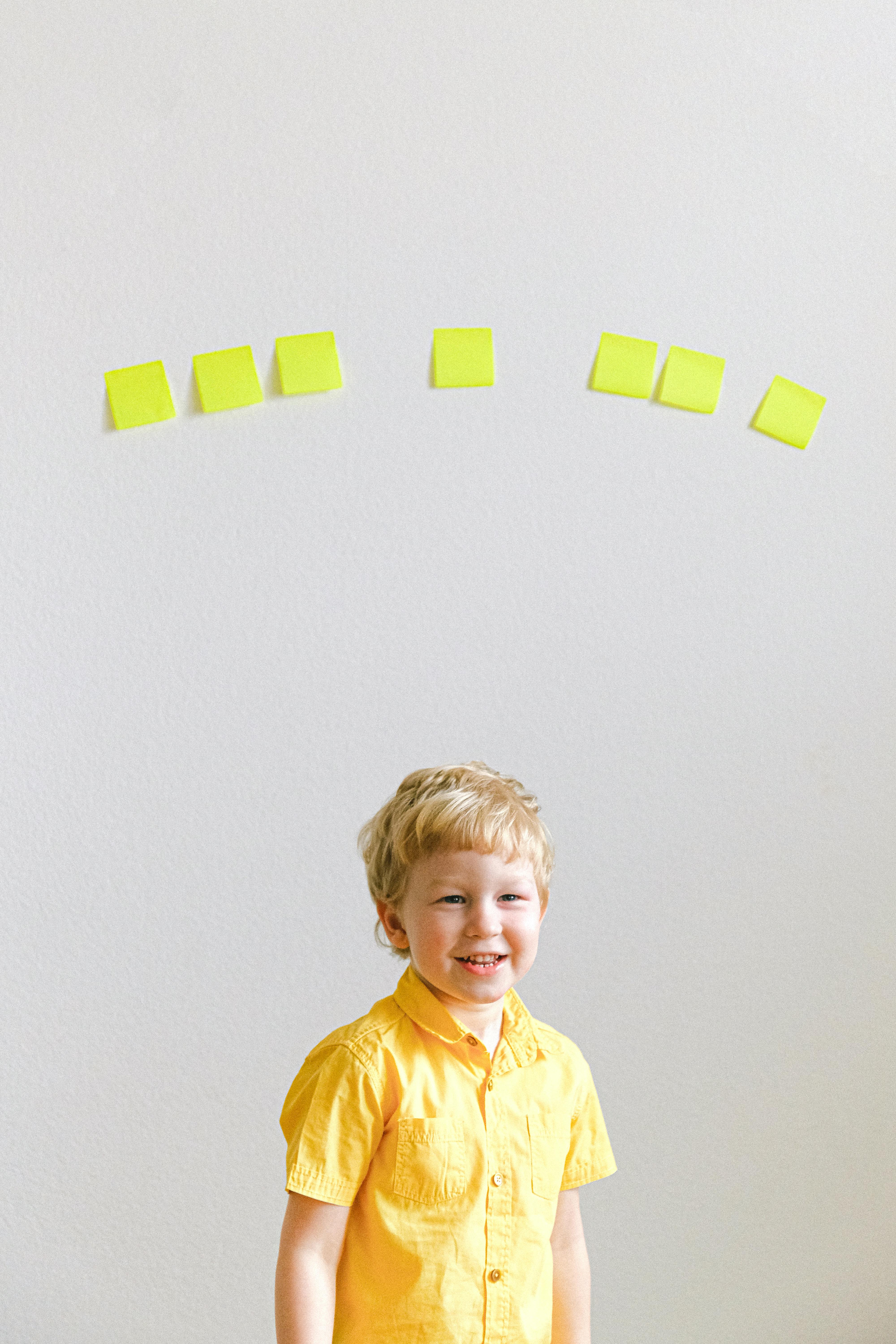 Adorable blonde child in yellow shirt smiling with sticky notes on the wall.