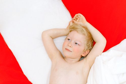 Free Adorable blond boy lying comfortably on a red and white bed, gazing thoughtfully. Stock Photo