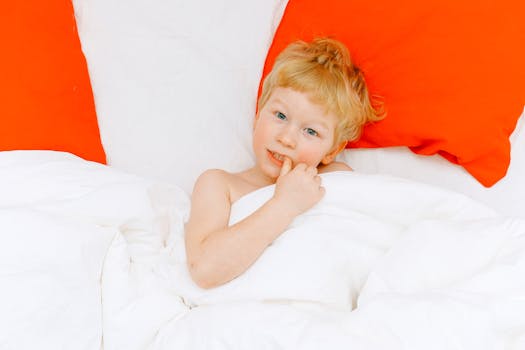Adorable blond child relaxing in bed with white sheets and red pillows, showcasing comfort and warmth.