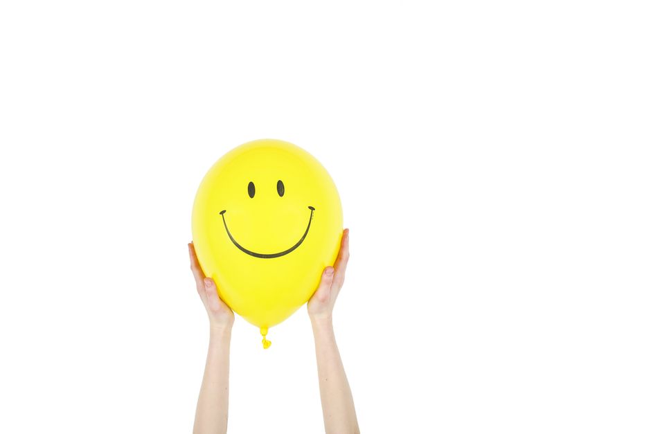 Fotomural Globo amarillo alegre con una cara sonriente sostenido por manos contra un fondo blanco.