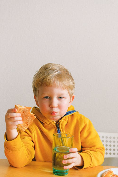 Boy Wearing Yellow Hoodie Eating Pizza And Holding A Glass Of Water