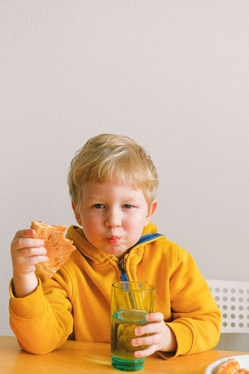 Free Young blond child in a yellow hoodie enjoying pizza and a drink at a table indoors. Cute and joyful moment. Stock Photo