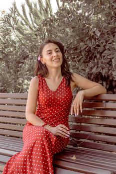 A woman in a red polka dot dress sitting on a park bench in Baku, Azerbaijan, smiling warmly.
