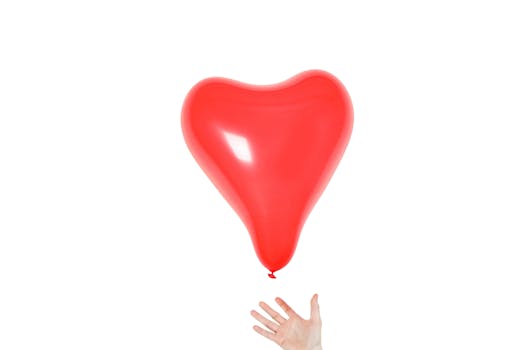 A vibrant red heart-shaped balloon floating with a hand reaching for it on a white background.