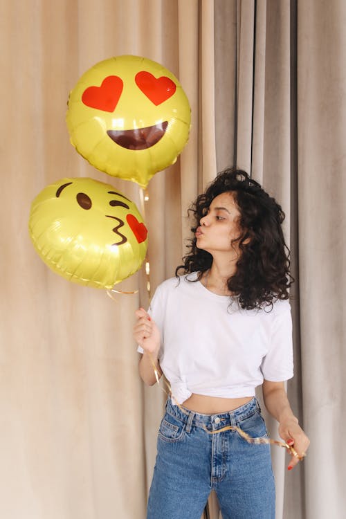 Free Positive young lady in white casual shirt and blue jeans pouting lips while looking at bright yellow balloons with emoticons in light room Stock Photo