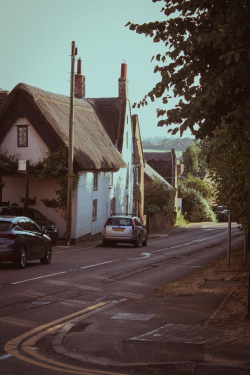 Free Scenic view of a quaint English village street with traditional thatched roof houses and parked cars. Stock Photo
