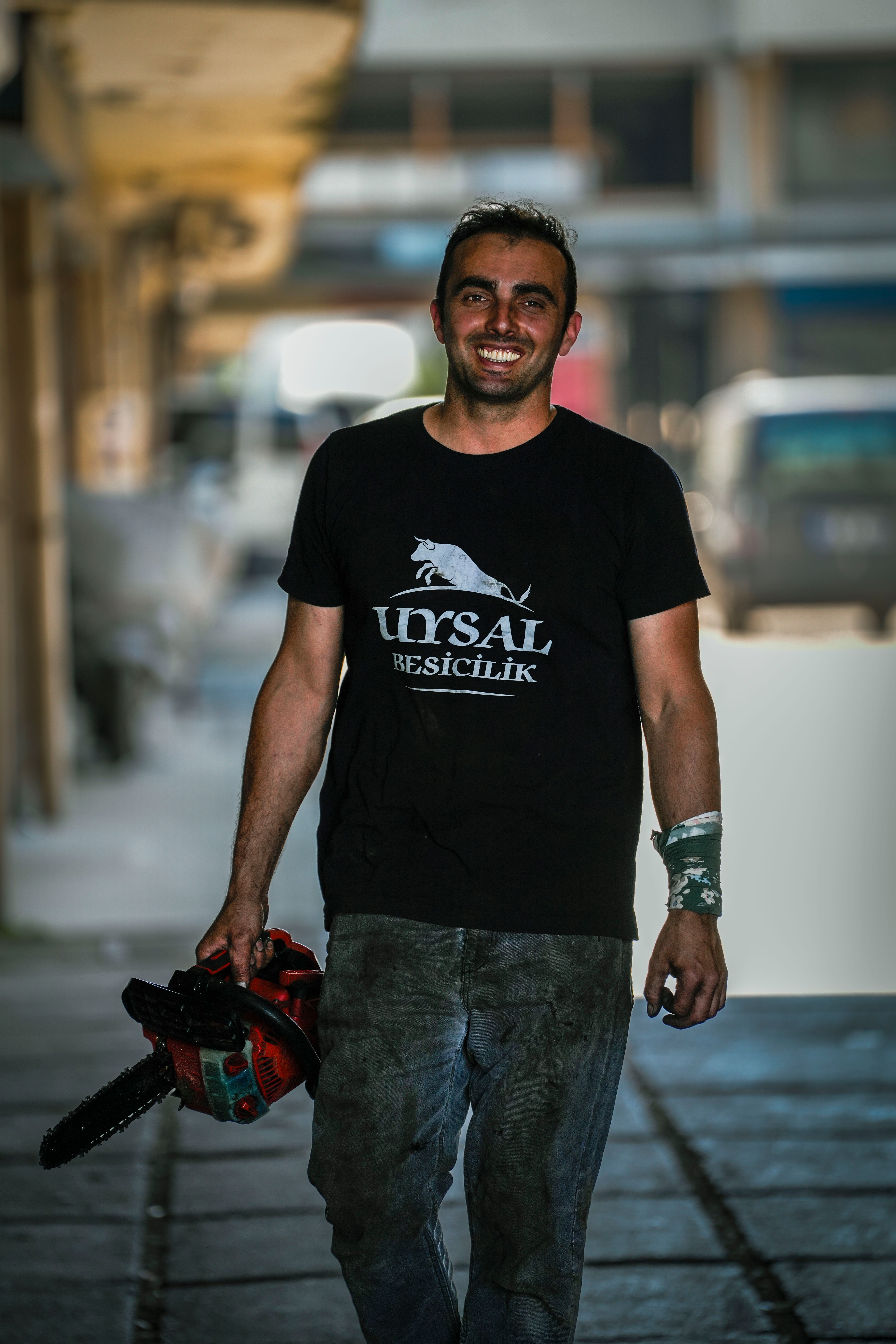 Man in a t-shirt smiling with a chainsaw in urban setting, conveying hard work and positivity.