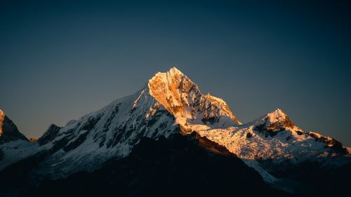 Free Golden sunlight illuminates a towering snow-covered mountain peak against a clear sky at sunrise. Stock Photo