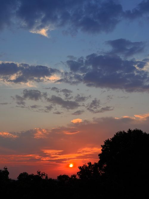 Free A beautiful sunset with vivid orange hues and clouds, casting shadows on silhouetted trees. Calm and peaceful. Stock Photo