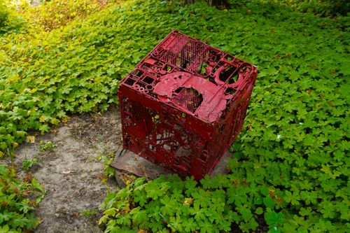 Free A striking red metal cube sculpture set amidst lush green foliage in an outdoor garden setting. Stock Photo