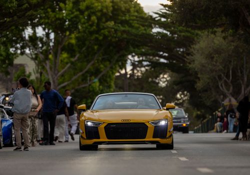 Yellow convertible sports car driving on a tree-lined street with people in the background.
