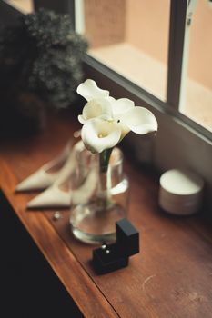A beautifully arranged wedding setup featuring a calla lily, bridal shoes, and a ring box by a window.