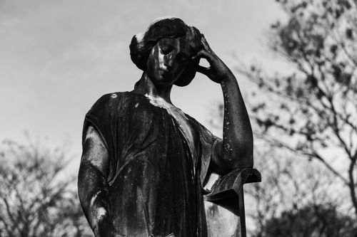 Free Black and white photo of a contemplative cemetery statue set against a backdrop of trees, symbolizing mourning. Stock Photo