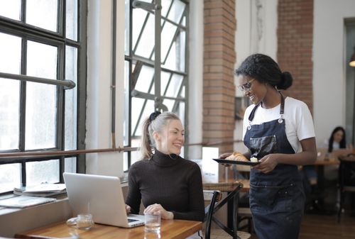 Free Friendly interaction in a café with a waitress serving a customer using a laptop. Bright and welcoming atmosphere. Stock Photo