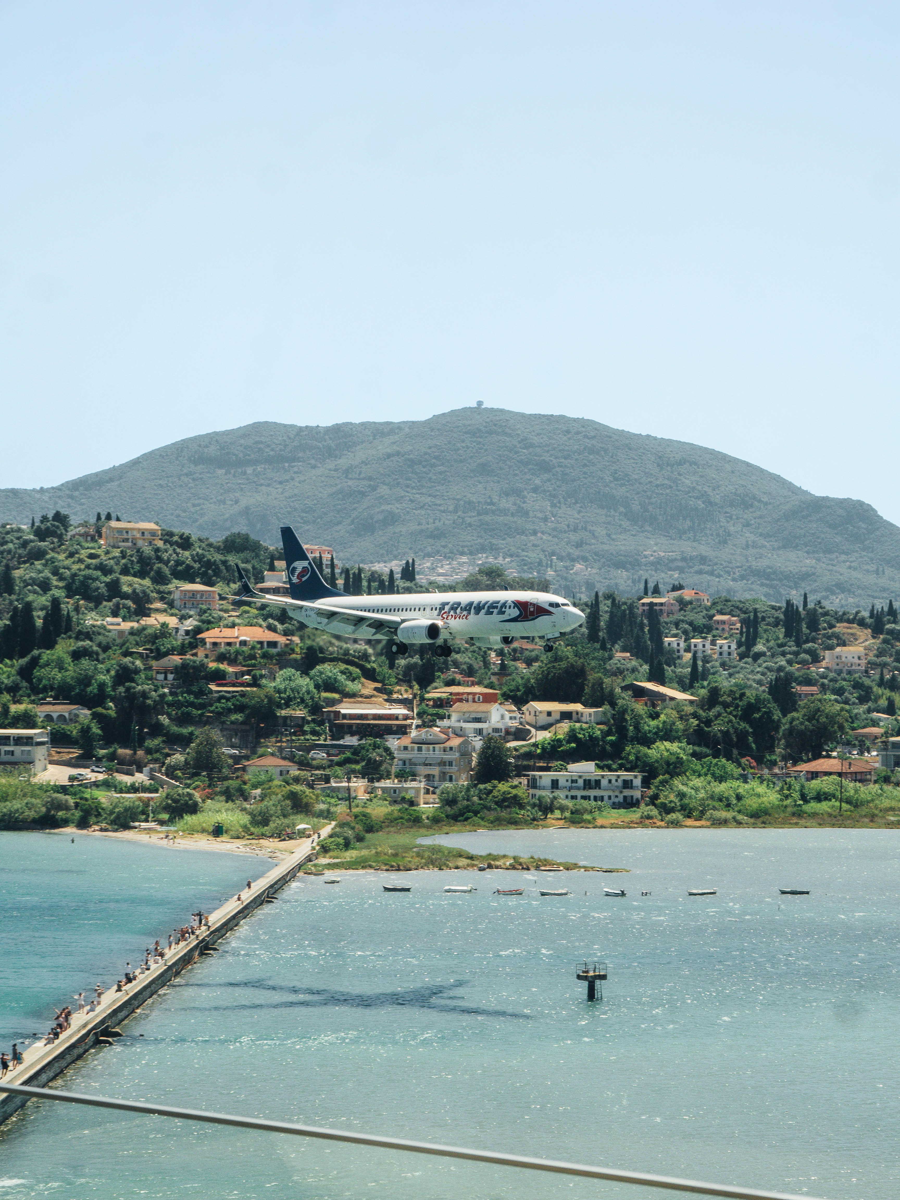 A commercial airplane flies over a vibrant coastal town with mountains in the background.