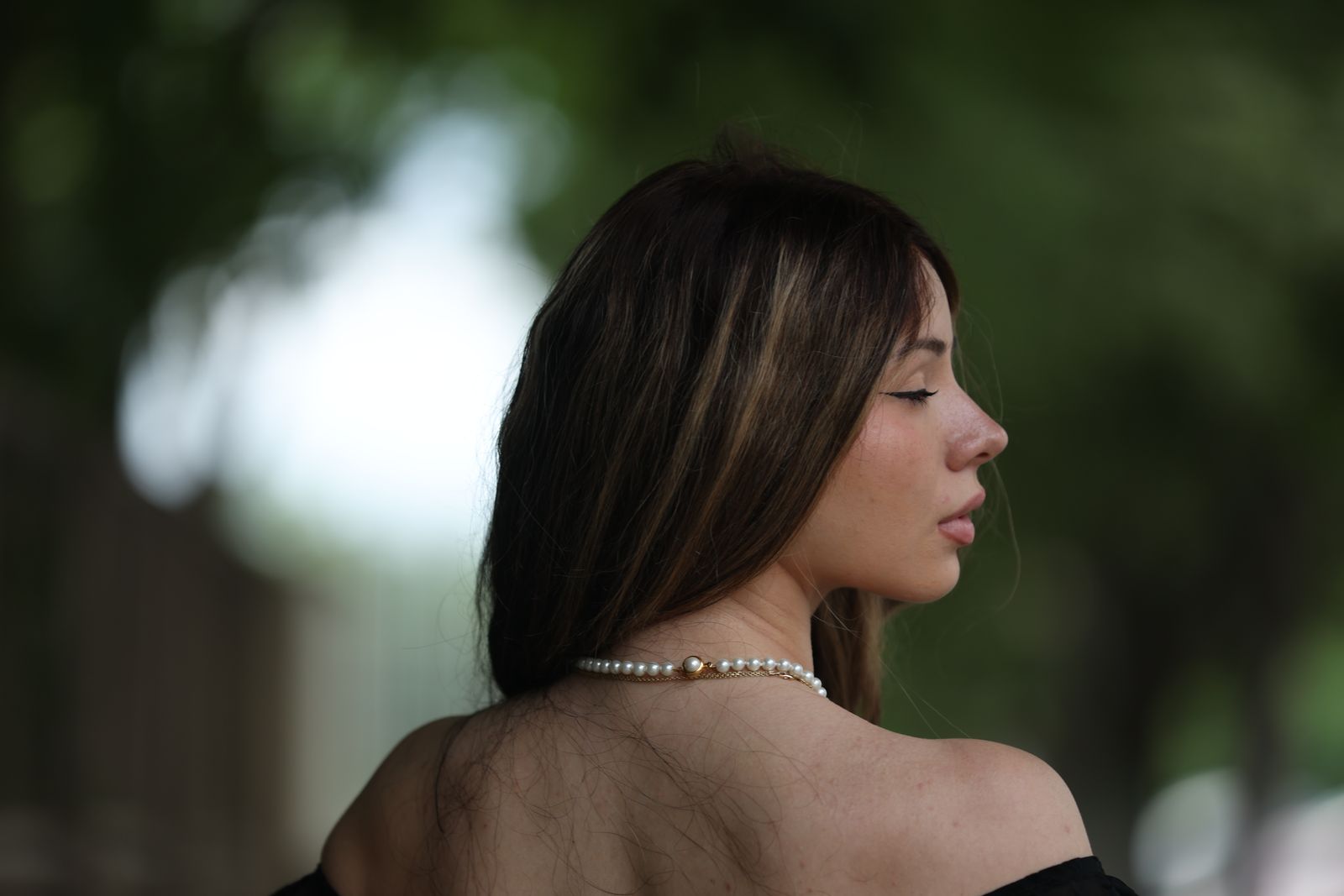 Woman with long brown hair in profile wearing a pearl necklace, bare shoulders, soft outdoor greenery behind