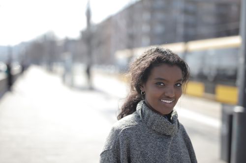 Free Young woman smiling on a sunny day in an urban setting, wearing a cozy sweater. Stock Photo