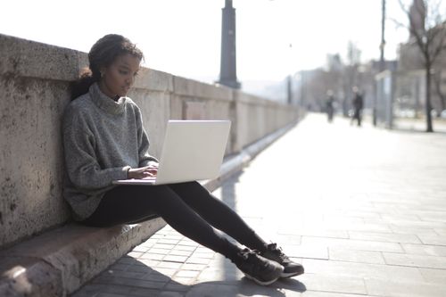 Free A young woman sits outdoors on a city street working remotely on her laptop. Stock Photo