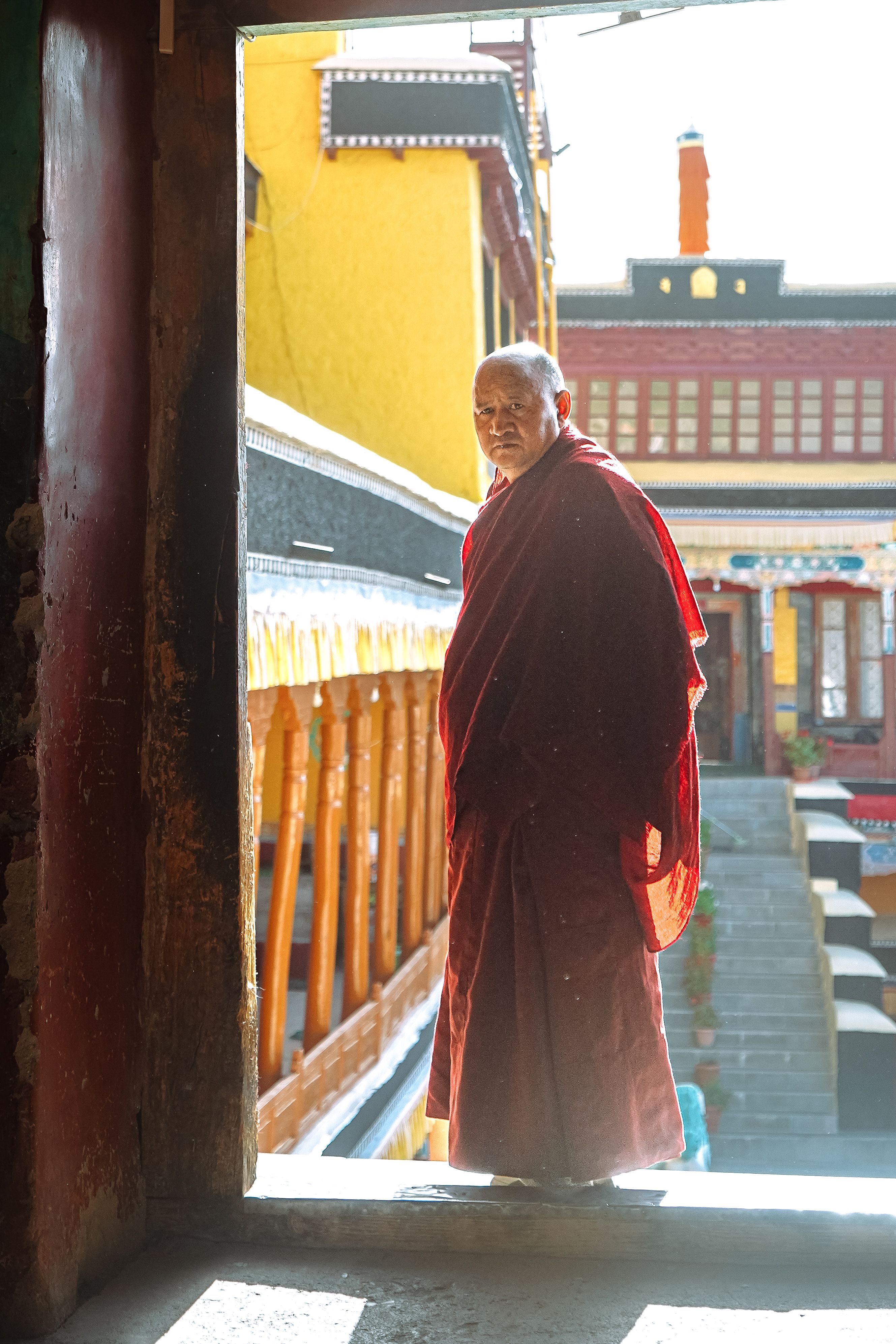 A monk in traditional attire stands in a sunlit corridor of an Indian monastery.