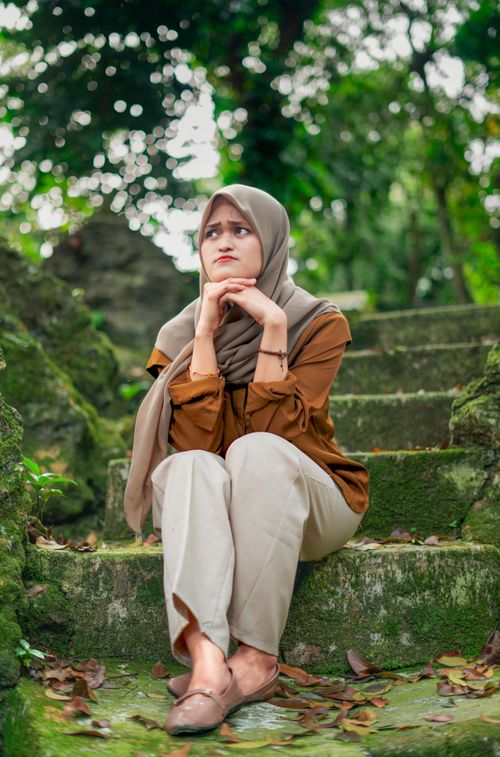 Free A contemplative woman in a hijab sits thoughtfully on moss-covered steps in a lush outdoor setting. Stock Photo