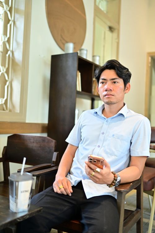Free Young man sitting casually in a modern indoor setting, holding a smartphone. Relaxed atmosphere. Stock Photo