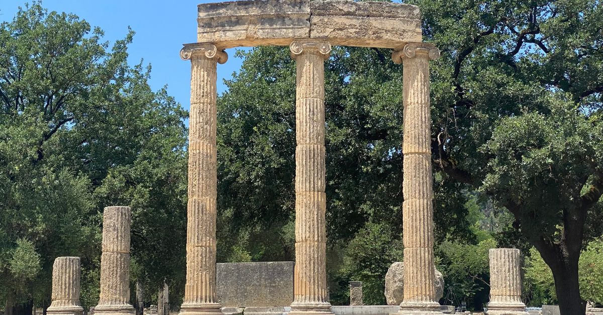 Explore the ancient Philippeion in Olympia, surrounded by lush greenery under a bright sky.