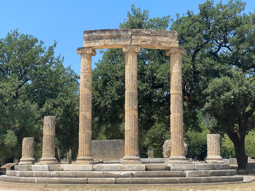 Explore the ancient Philippeion in Olympia, surrounded by lush greenery under a bright sky.