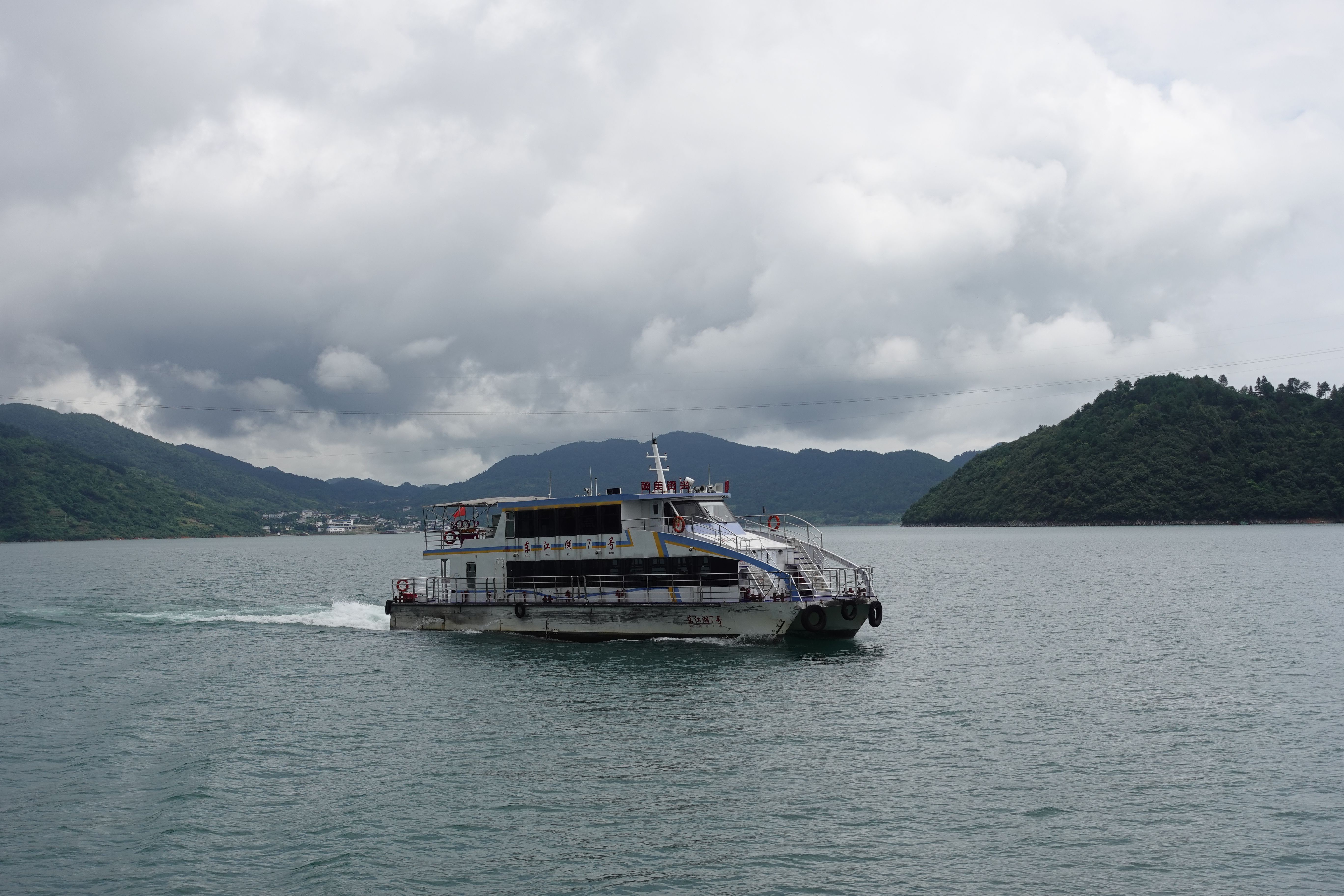 Passenger ferry on a serene lake with lush hills and cloudy skies.