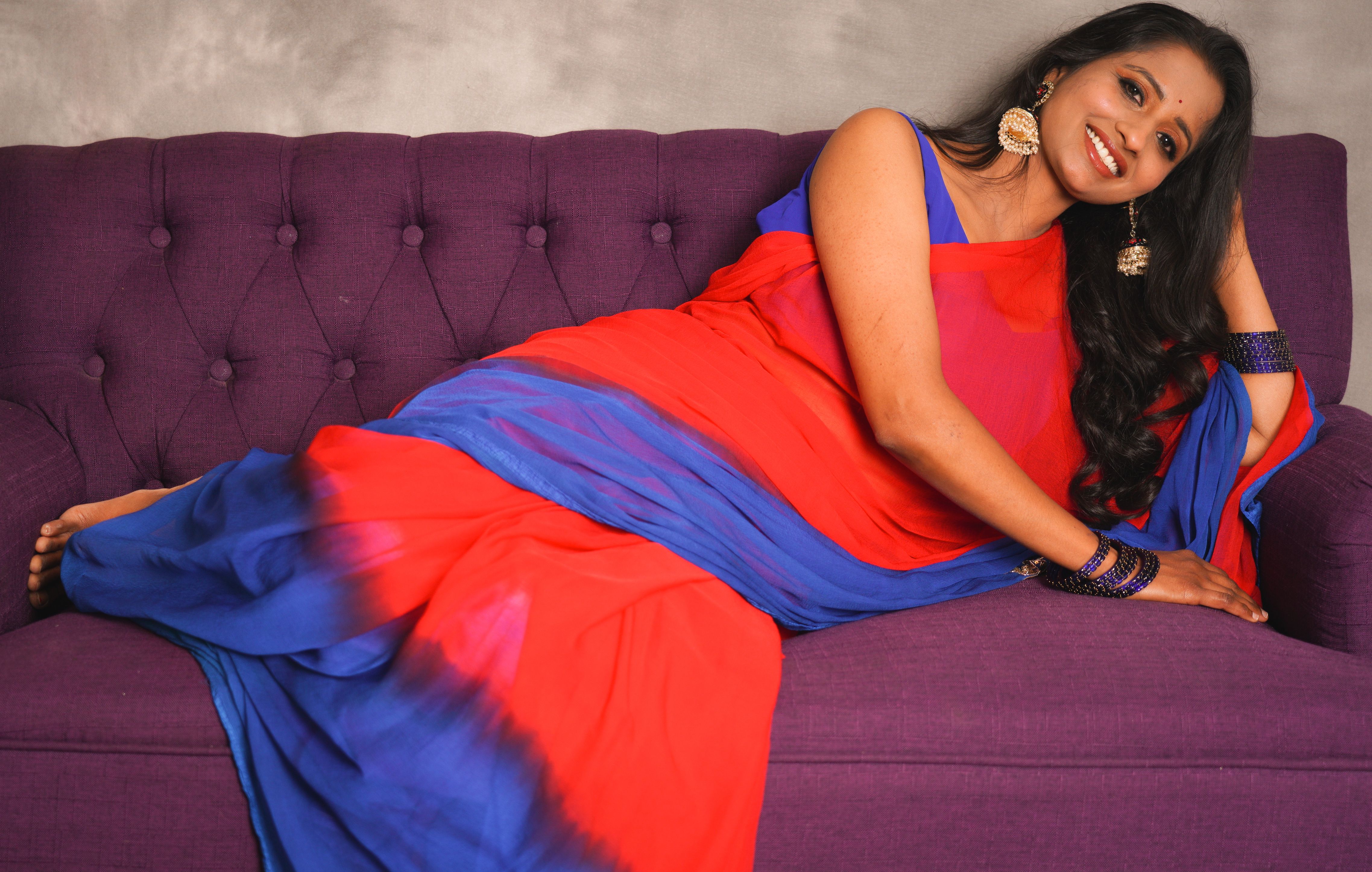 A woman in a red and blue sari reclines on a purple sofa, smiling warmly.